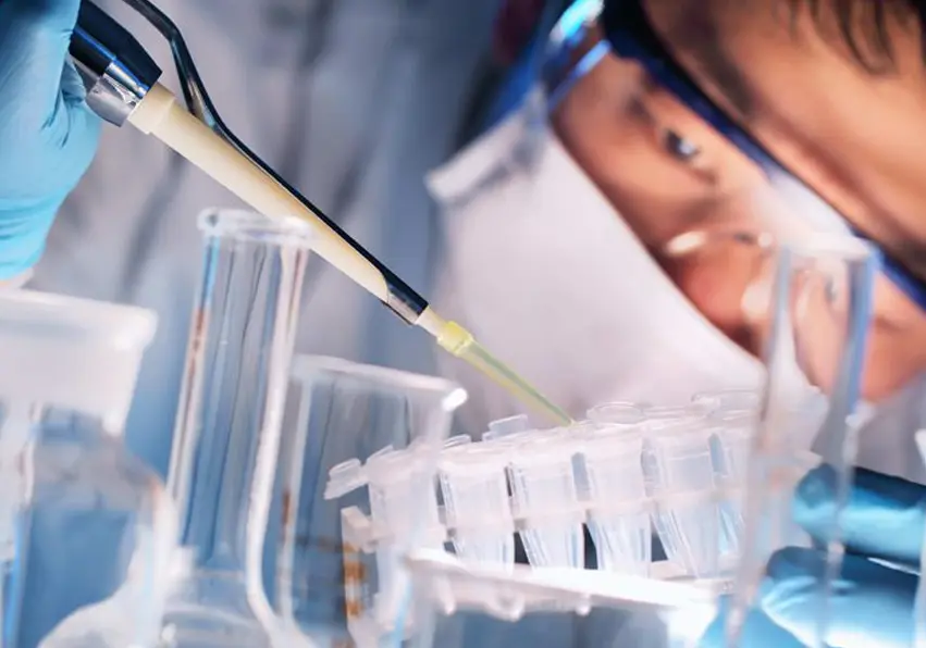 A person in lab coat and goggles working with beakers.