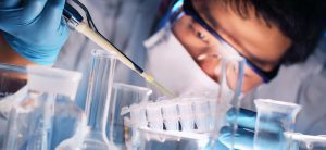 A person in lab coat and goggles working with beakers.