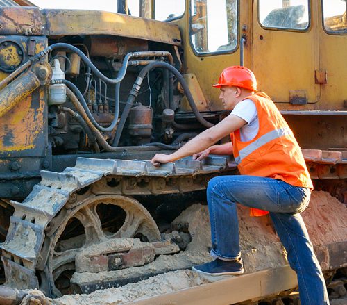 A man in an orange vest is on the back of a yellow tractor.